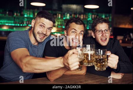 Klopfen Gläser. Drei sport Fans in einer Bar beobachten Fußball. Mit einem Bier in der Hand Stockfoto