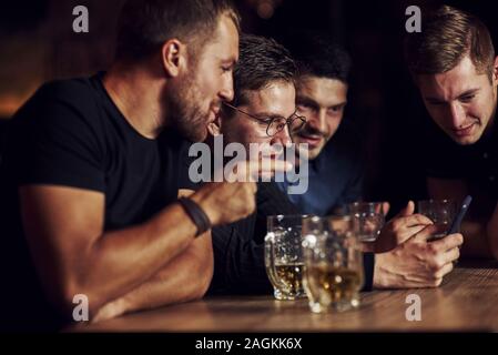 Lustige Inhalte auf dem Smartphone. Drei Freunde in der Kneipe ruht mit einem Bier in der Hand. In Gespräch Stockfoto