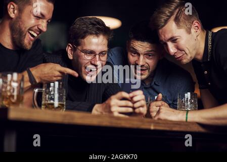 Lustige Inhalte auf dem Smartphone. Drei Freunde in der Kneipe ruht mit einem Bier in der Hand. In Gespräch Stockfoto