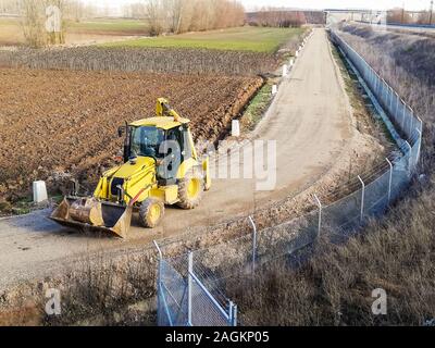 Ein Baggerlader auf einem Feldweg zwischen den Kulturen neben einer Eisenbahnlinie Zaun. Bauweise Stockfoto