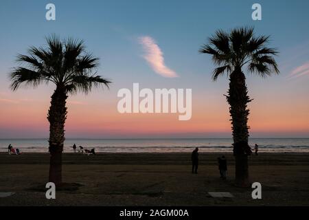 Larnaca, Zypern - 2. Januar 2018: Menschen bei Finikoudes Strand, einem Spaziergang bei Sonnenuntergang. Palm Tree Silhouetten und bunte Himmel im Blick. Stockfoto