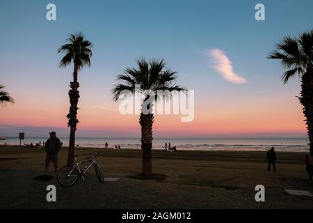 Larnaca, Zypern - 2. Januar 2018: Menschen bei Finikoudes Strand, einem Spaziergang bei Sonnenuntergang. Palm Tree Silhouetten und bunte Himmel im Blick. Stockfoto