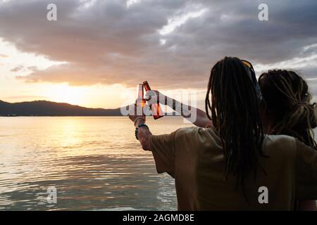 Ansicht der Rückseite des Paar genießen Sie einen wunderschönen Sonnenuntergang und Bierflaschen klirren, wenn die Ausgaben romantischen Abend am Strand. Stockfoto