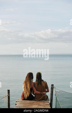 Ansicht der Rückseite des umarmen Paar sitzt auf schmale Holzbrücke und Suchen im schönen ruhigen Meer Stockfoto