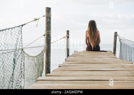 Nachdenkliche junge Frau mit dreadlock sitzt auf schmalen handgefertigten Holz- pier und auf schönen ruhigen Ozean Stockfoto