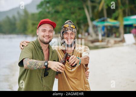 Glücklich lächelnde Freunde umarmen und klirrenden Flaschen Bier im Stehen am Strand. Stockfoto