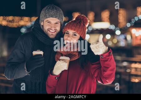 Foto von zwei Menschen mit heißem Tee trinken in Händen feiern x-mas Eve in der Magie im freien Atmosphäre heben Daumen hoch tragen warme Mäntel Stockfoto