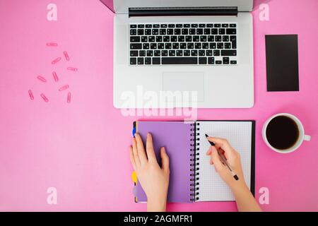 Überblick über weibliche Hände in einem offenen Notepad schreiben. Kaffee, Laptop und Stift neben. Flach gestaltete moderne Büroflächen. Stockfoto