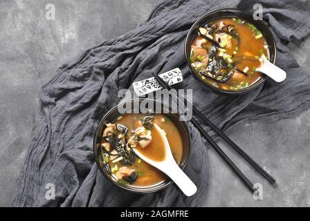 Japanische Miso-Suppe mit frischem Thunfisch, getrockneten Algen, Tofu, Shiitake getrockneten Pilzen in einer schwarzen Schüssel auf grauem Hintergrund Stockfoto