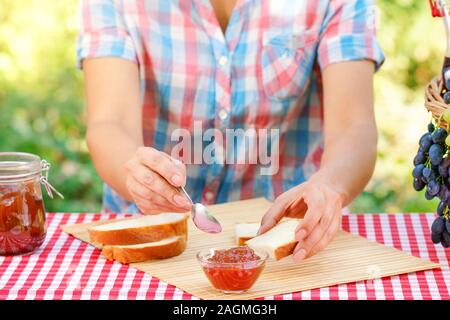 Frau in Plaid Shirt Brotaufstriche Marmelade auf Brot. Rot Tischdecke, Picknickkorb, Trauben. Sonnigen Tag. Picknick Konzept Stockfoto