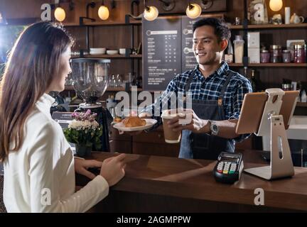 Portrait von asiatischen Barista halten nehmen Sie Kaffee und Croissant Cup und dem Kunden in Cafe, das mit anderen Barista im Hintergrund arbeiten Stockfoto