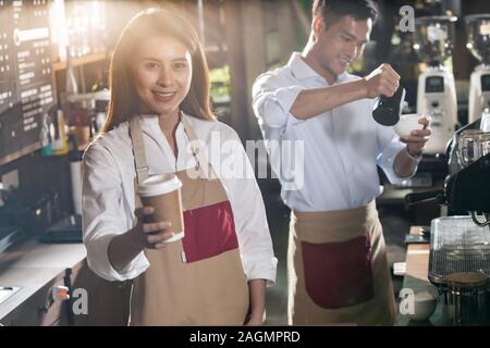 Portrait von asiatischen Barista halten nehmen Sie Kaffee Tasse und an Kunden im Cafe, das mit anderen Barista im Hintergrund arbeiten Stockfoto
