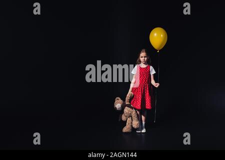 Trauriges Kind mit gelben Ballon und Teddybären an Kamera isoliert auf Schwarz auf der Suche Stockfoto