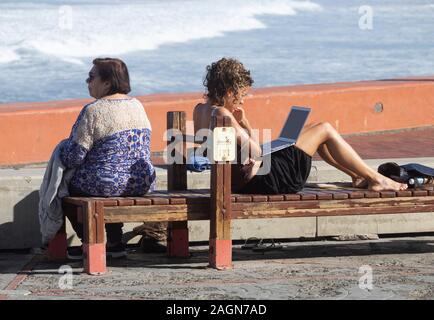 Jüngere Frau am Strand sitzen auf gym Sitzbank bei Laptop suchen, während ältere Frau auf das Meer blickt. Stockfoto