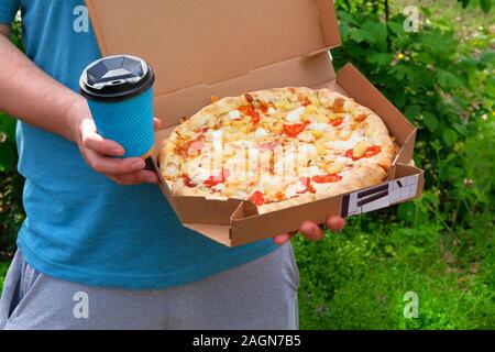 Mann im blauen T-Shirt ist Holding Box mit lecker Pizza und Pappbecher mit Getränk, die Ansicht zu schließen. Italienische Küche. Snack an einem sonnigen Sommertag. Stockfoto