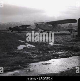 1960, historische, kleine 2-Sitzer flächenflugzeug Propeller- Flugzeugen sitzen auf unebenem Gelände in der Nähe von Flughafen Anchorage, Anchorage, Alaska, USA, mit der Chugach Mountains im Hintergrund. Stockfoto