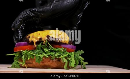 Handwerk Burger ist Kochen auf schwarzem Hintergrund. Bestehen: sauce, Rucola, Tomaten, Zwiebeln, Speck, Johannisbeeren Sauce, ricotta Käse, Brötchen und Fleisch. Stockfoto