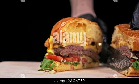 Schneiden Handwerk Burger ist Kochen auf schwarzem Hintergrund. Bestehen: sauce, Rucola, Tomaten, roten Zwiebeln Pommes, Gurken, Käse, Brötchen und Fleisch. Stockfoto