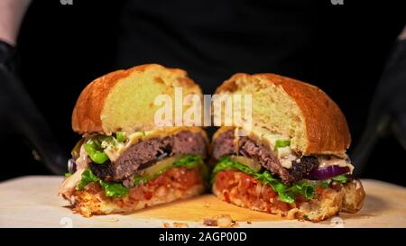 Schneiden Handwerk Burger ist Kochen auf schwarzem Hintergrund. Bestehen salsa Sauce, Salat, rote Zwiebel, Gurke, Käse, Chili, Paprika, Brötchen, marmoriertes Fleisch Rindfleisch. Stockfoto