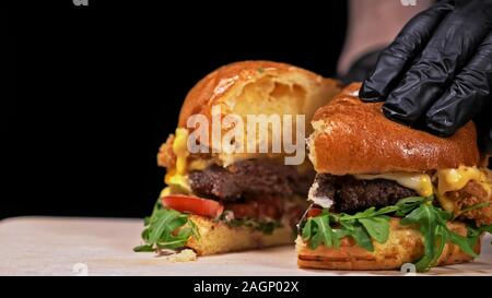 Schneiden Handwerk Burger ist Kochen auf schwarzem Hintergrund. Bestehen: sauce, Rucola, Tomaten, roten Zwiebeln Pommes, Gurken, Käse, Brötchen und Fleisch. Stockfoto