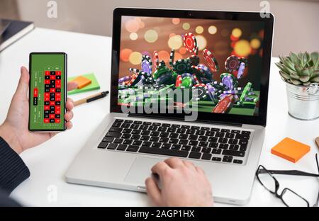 Online Glücksspiel, wetten. Poker chips, casino roulette auf Laptop und Handy Bilder. Mann bei der Arbeit mit einem Computer und einem Smartphone, offi Stockfoto