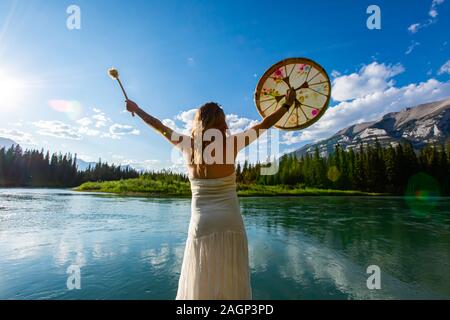 Traditionelle einheimische Kultur trifft Natur als achtsam Frau Tragen eines weißen Sommerkleid hält die heilige Trommel und die Wendetrommel durch einen Fluss Stockfoto