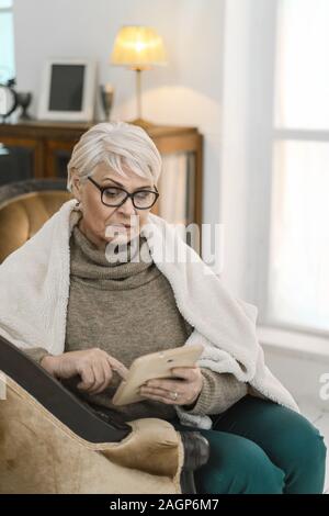 Thougtful alte Dame in Gläsern ist entspannend auf Ihre komfortable Wohnung. Sie sitzt auf dem Leder Sessel und mit Ihrem Tablet-PC. Die Frau ist Gewickelt Stockfoto