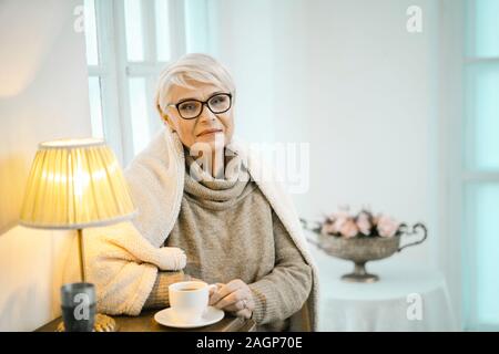 Alte Frau in einem Wollpullover und eine karierte auf Ihren Schultern steht in der Nähe der Schränke. Sie hält eine Tasse Tee. Die Leuchte wird eingeschaltet. Vase W Stockfoto