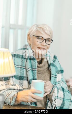 Ältere Frau Brille ist verpackt in einem karierten Plaid zum Wohlfühlen. Granny ist Tee trinken. Eingeschaltete Leuchte ist neben ihr. Stockfoto