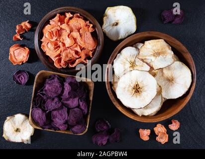 Karotte, rote Bete und Apfel Chips in eine hölzerne Schüssel auf einen schwarzen Stein getrocknet. Organische natürliche Nahrung. Ansicht von oben, flach. Stockfoto