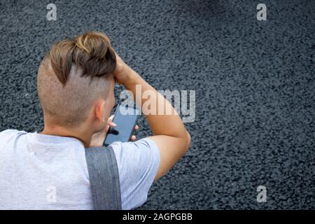 Rückseite der Kopf rasiert mit Pferdeschwanz junger Mann mit Smartphone gegen grauen Asphalt Stockfoto