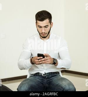 Ein junger Mann mit Bart in einem weißen Hemd arbeitet mit einem Smartphone beim Sitzen auf einem Stuhl in einem hellen Zimmer Stockfoto