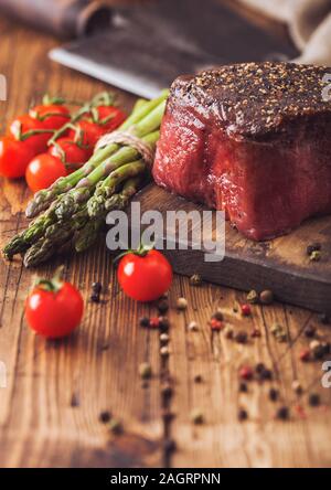 Ein Stück rohes Rindfleisch Oberschale mit Salz und Pfeffer auf Holz Schneidbrett mit Tomaten, Knoblauch und Spargelspitzen auf Holz Küche Tisch. Stockfoto