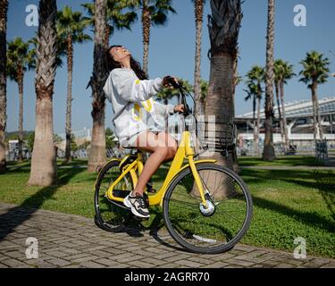 Ein junges Mädchen in einem Hoodie auf einem gelben Fahrrad mit einem Lächeln reitet entlang einer Palm Avenue. Sommer Spaß Glück Freude Stockfoto