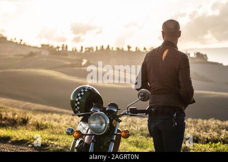 Mann stand neben seinem Motorrad entlang einer Landstraße auf Hügel Landschaft bei Sonnenuntergang. Toskana, Italien. Stockfoto