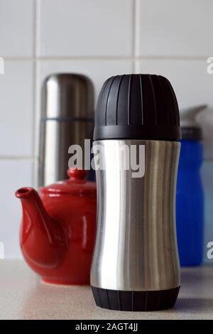Zwei geschlossene Metall heißen Getränk Flaschen. Ein roter Tee pot. Eine blaue Flasche Wasser. Weiße Fliesen- Hintergrund. Stockfoto
