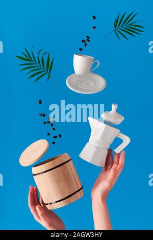 Trendy Levitation. Fliegen Linie der Kaffeebohnen zwischen Keramik Kaffeemaschine und Espresso Tasse mit Untertasse. Blue mint Hintergrund mit natürlichen Palm verlassen Stockfoto