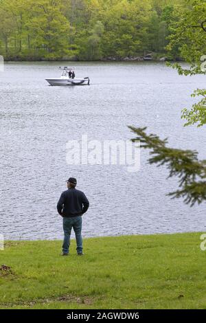 Neue Preston, CT USA. Mai 2016. Mann am Ufer des Lake Waramaug als eine Polizeistreife Motorboot vorbei im Hintergrund. Stockfoto