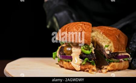 Schneiden Handwerk Burger ist Kochen auf schwarzem Hintergrund. Bestehen salsa Sauce, Salat, rote Zwiebel, Gurke, Käse, Chili, Paprika, Brötchen, marmoriertes Fleisch Rindfleisch. Stockfoto