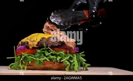 Handwerk Burger ist Kochen auf schwarzem Hintergrund. Bestehen: sauce, Rucola, Tomaten, Zwiebeln, Speck, Johannisbeeren Sauce, ricotta Käse, Brötchen und Fleisch. Stockfoto