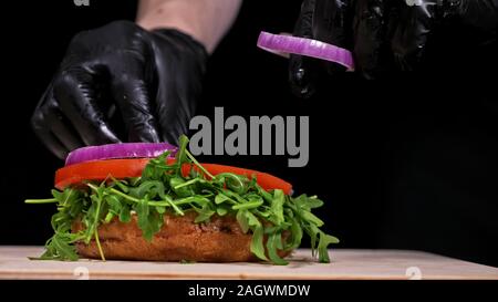 Handwerk Burger ist Kochen auf schwarzem Hintergrund. Bestehen: sauce, Rucola, Tomaten, Zwiebeln, Speck, Johannisbeeren Sauce, ricotta Käse, Brötchen und Fleisch. Stockfoto