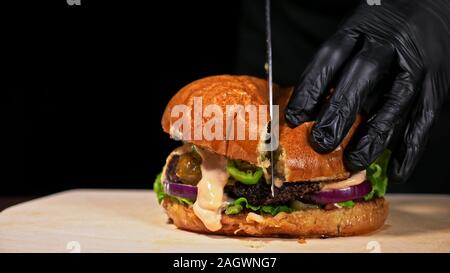 Schneiden Handwerk Burger ist Kochen auf schwarzem Hintergrund. Bestehen salsa Sauce, Salat, rote Zwiebel, Gurke, Käse, Chili, Paprika, Brötchen, marmoriertes Fleisch Rindfleisch. Stockfoto