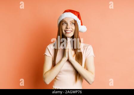 Fröhliche kaukasischen Sommersprossige rothaarige Frau in Santa hat zu beten Geste, Palmen zusammen halten, breit grinsend, über Coral Hintergrund isoliert Stockfoto