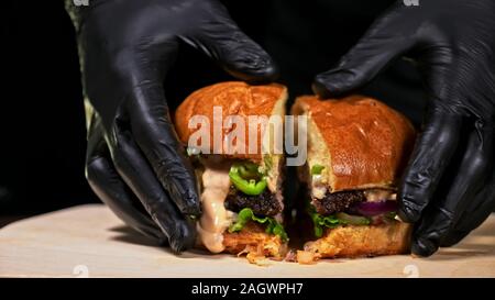 Schneiden Handwerk Burger ist Kochen auf schwarzem Hintergrund. Bestehen salsa Sauce, Salat, rote Zwiebel, Gurke, Käse, Chili, Paprika, Brötchen, marmoriertes Fleisch Rindfleisch. Stockfoto