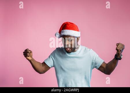 Wütend, wütend afrikanischer Mann ballte die Fäuste und Zähne, Hände, tragen t-shirt, Santa Hut, Pläne für Weihnachten nicht wahr, auf Rosa posing b Stockfoto