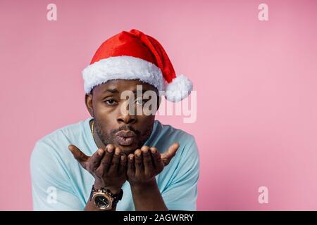Closeup Schuß von Stattlichen afro-amerikanische Mann mit Luft ausblasen Kiss über beide Handflächen, tragen, Santa Hut, Armbanduhr, weisses T-Shirt, mit dem Ausdruck der Zuneigung und l Stockfoto