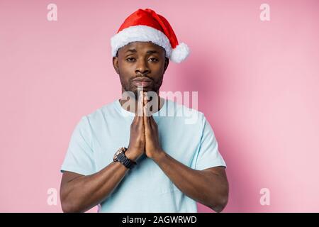 Ernsthafte dunkelhäutige Menschen zu Gott beten, Palmen zusammen halten, tragen, Santa Hut, über rosa Hintergrund mit Kopie Raum isoliert. Glaube, Hoffnung, religi Stockfoto