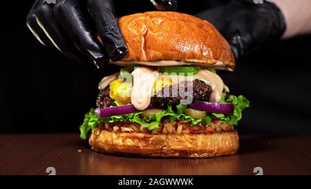 Handwerk Burger ist Kochen auf schwarzem Hintergrund. Bestehen: salsa Sauce, Salat, rote Zwiebel, Gurke, Käse, Chili, Paprika, Brötchen, marmoriertes Fleisch Rindfleisch. Stockfoto