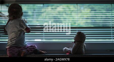 Kleinkind Mädchen schauen aus dem Fenster, durch abgesenkt Jalousien mit ihrem Teddybär neben ihr sitzt in einem konzeptionellen Bild. Stockfoto