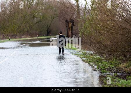 Earith, UK. 22 Dez, 2019. Eine Frau geht durch eine überflutete Straße wie der Fluss Great Ouse Überschwemmungen nach mehreren Tagen Regen. Die Umweltagentur hat zahlreiche Hochwasserwarnungen in Großbritannien ausgestellt als der längere Zeit der nassen Winter Wetter hat große Ansammlungen von Wasser im Fluß-Systeme verursacht. Die Ouse Washes sind Teil der Moorlandzone drainage System und soll den von der Überschwemmung mit Wasser in die flache Landschaft zu lösen. Credit: Julian Eales/Alamy leben Nachrichten Stockfoto